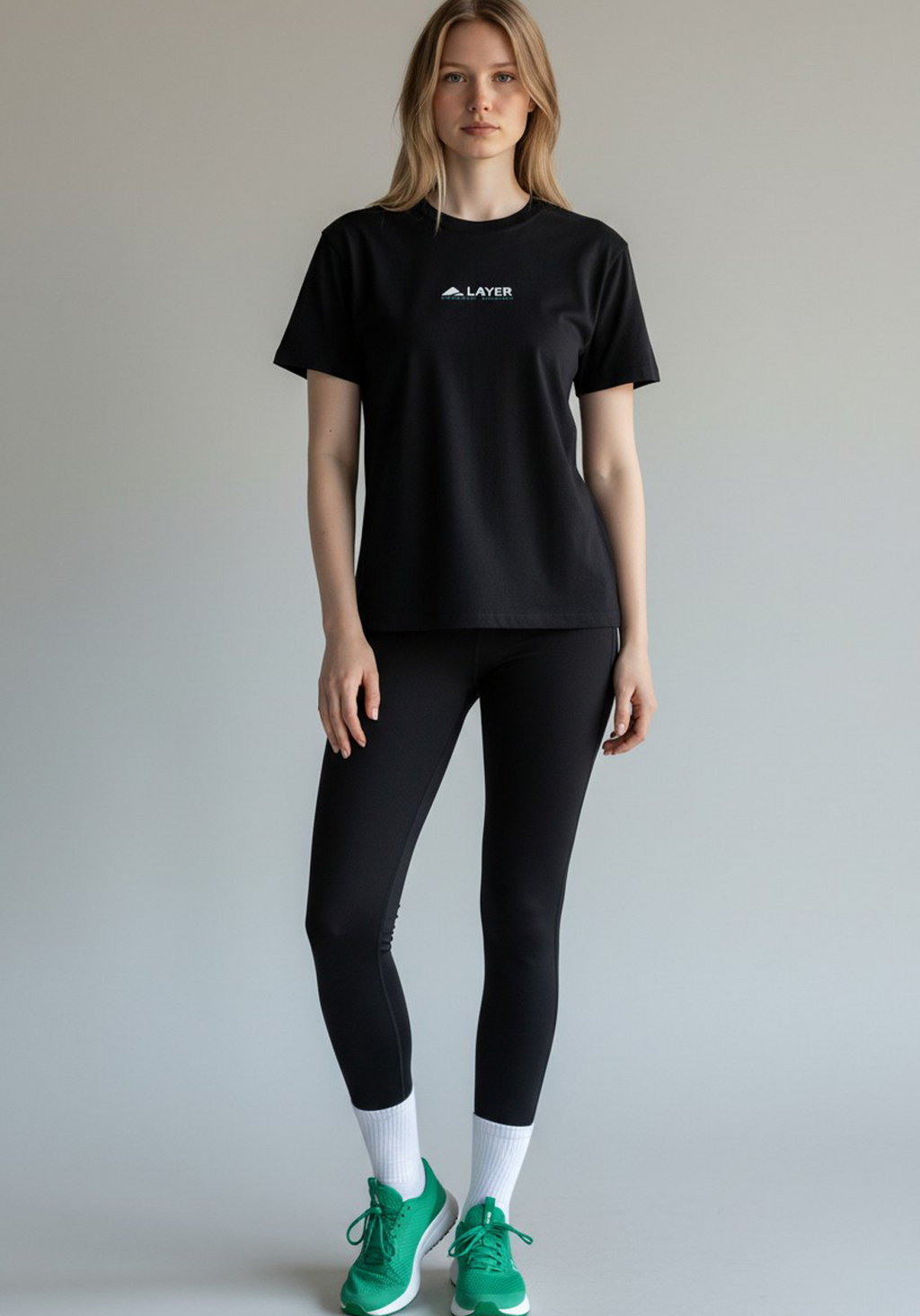 Full-length view of a woman wearing a black Layer Performance Sport Signature Tee, black leggings, white socks, and green trainers.