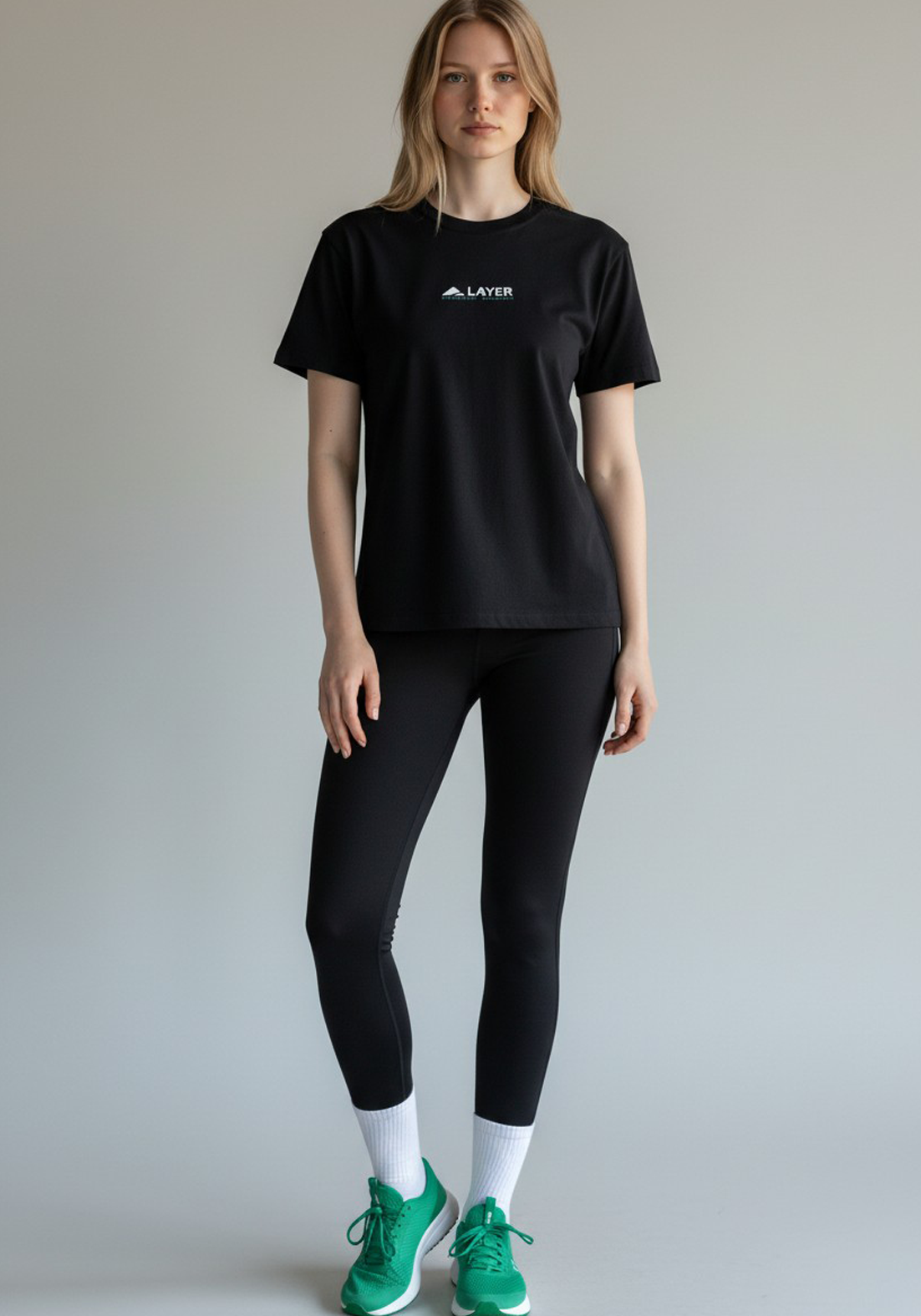 Full-length view of a woman wearing a black Layer Performance Sport Signature Tee, black leggings, white socks, and green trainers.