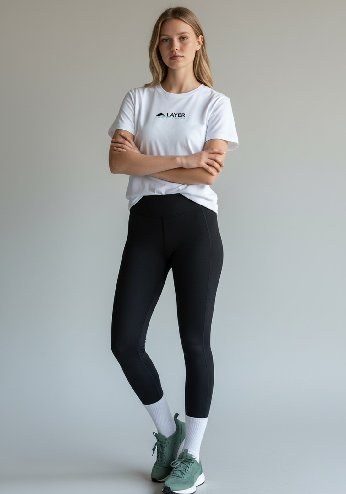 Woman wearing a white Layer Performance Sport Signature Tee and black leggings, standing with her arms crossed.