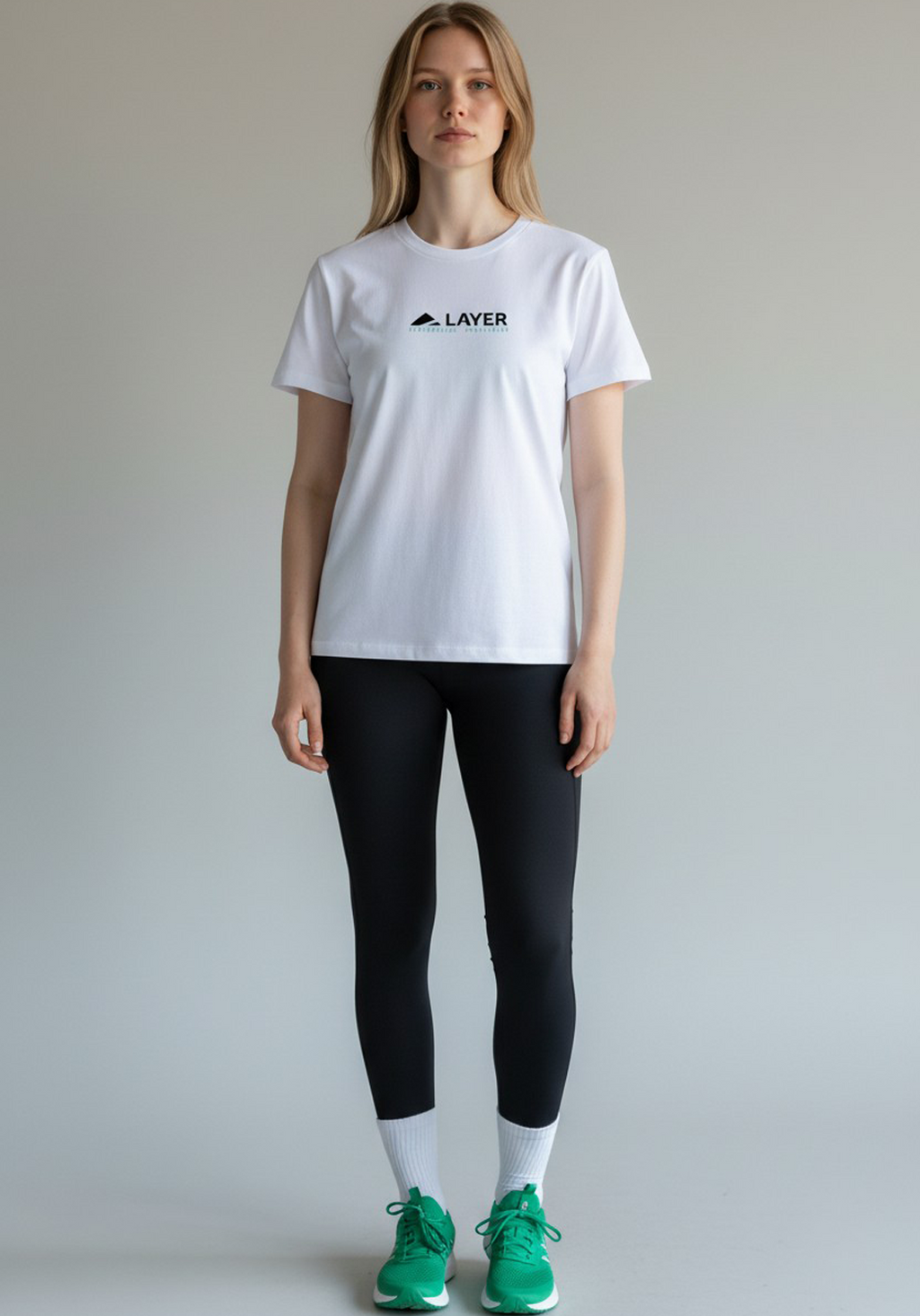 Woman standing full-length in a white Layer Performance Sport Signature Tee, black leggings, white socks, and green trainers.