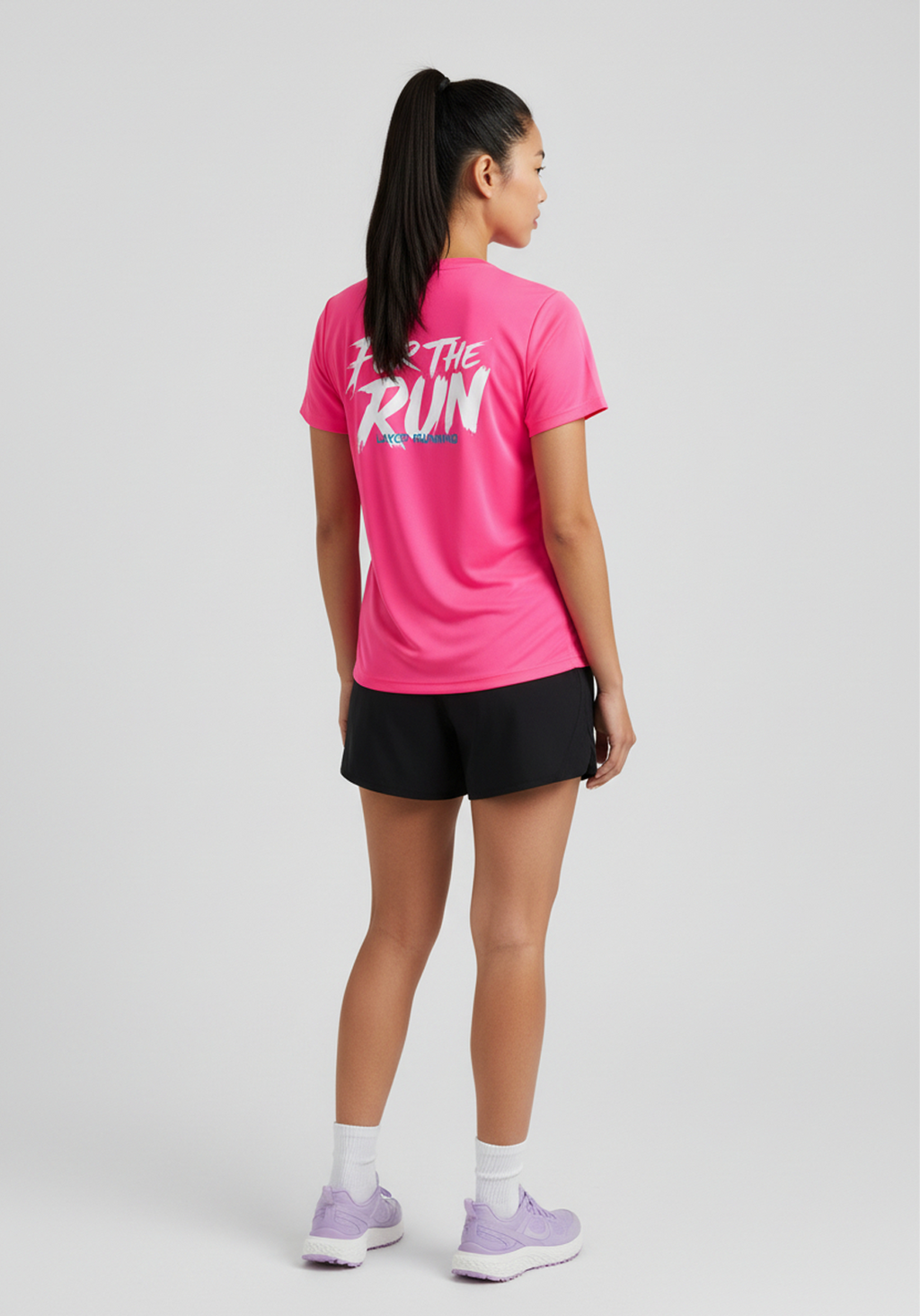 Back view of woman wearing pink Layer Women’s FTR Graphic Running Tee with ‘For The Run’ graphic, paired with black running shorts.