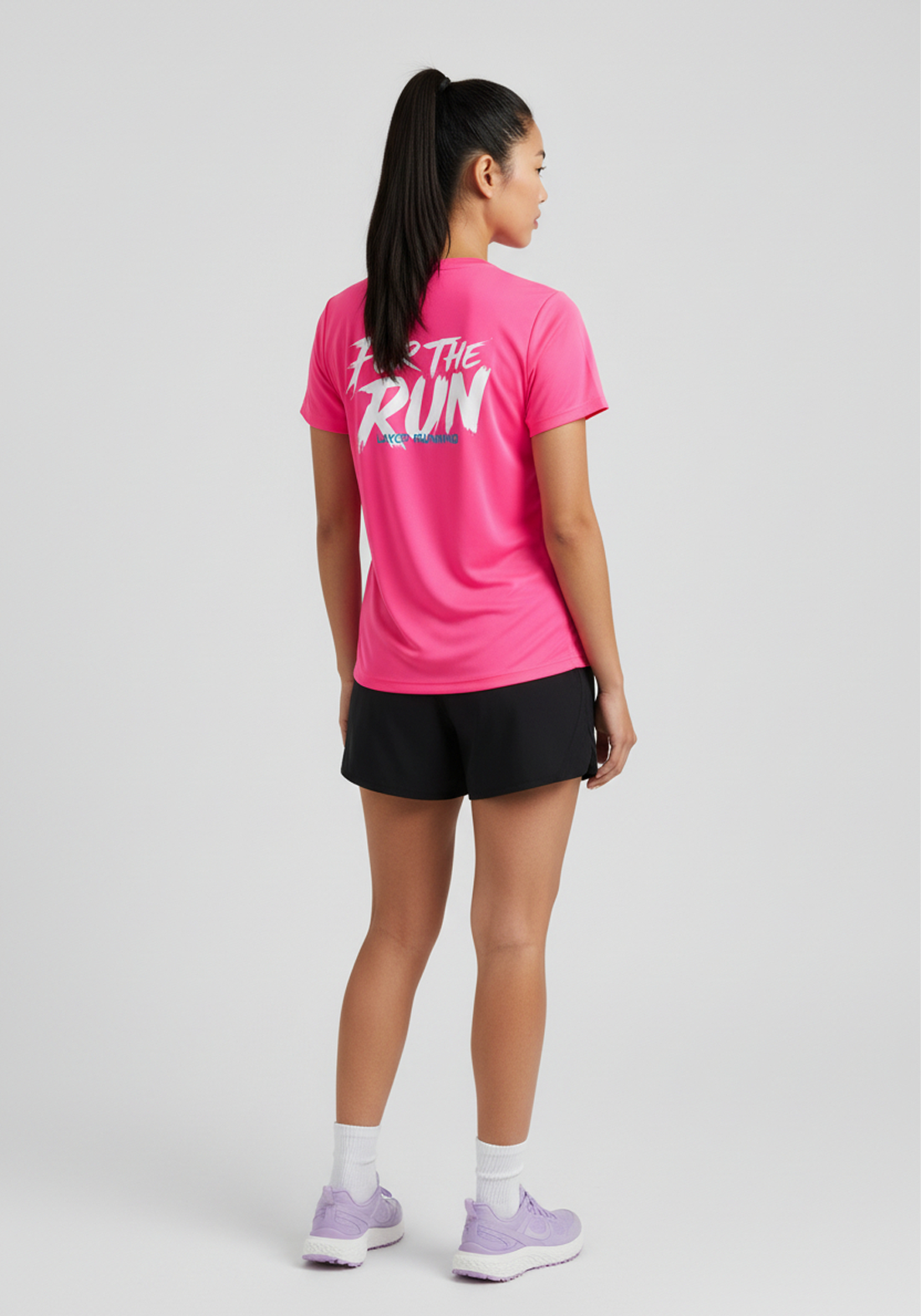 Back view of woman wearing pink Layer Women’s FTR Graphic Running Tee with ‘For The Run’ graphic, paired with black running shorts.
